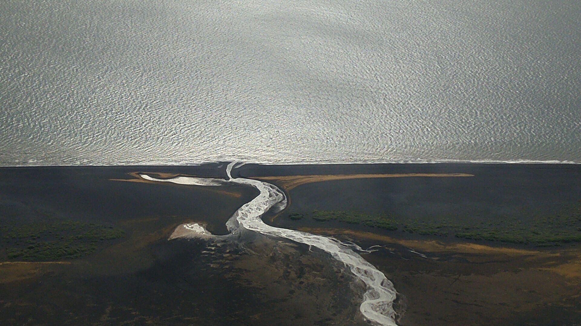 An aerial photo of a silver ribbon of a river, winding through a black beach towards a silver-looking ocean.