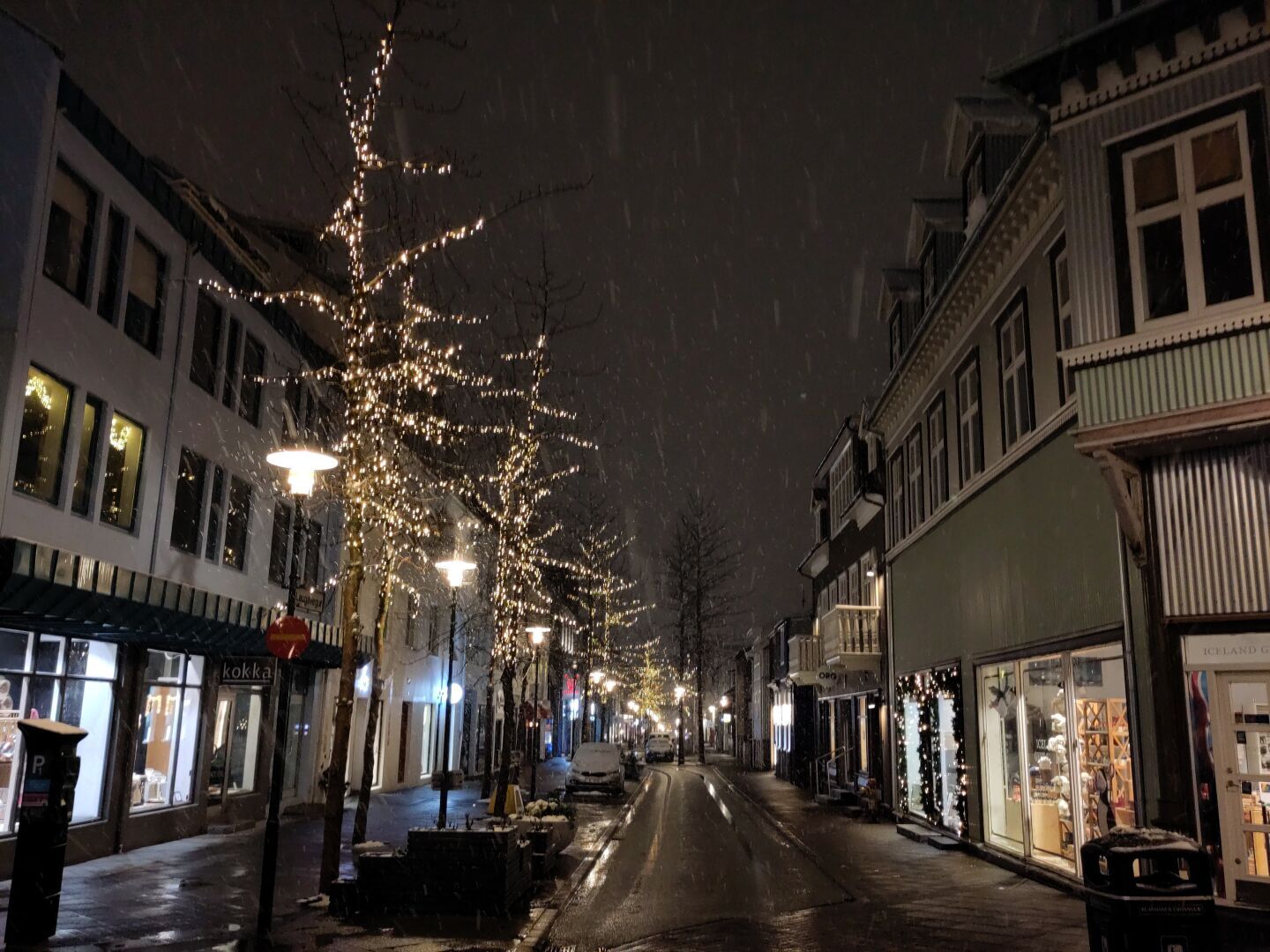 An empty narrow street with wide sidewalks and store fronts at night. Parked cars covered in snow. Trees lit up with white Christmas lights. Snow falling.