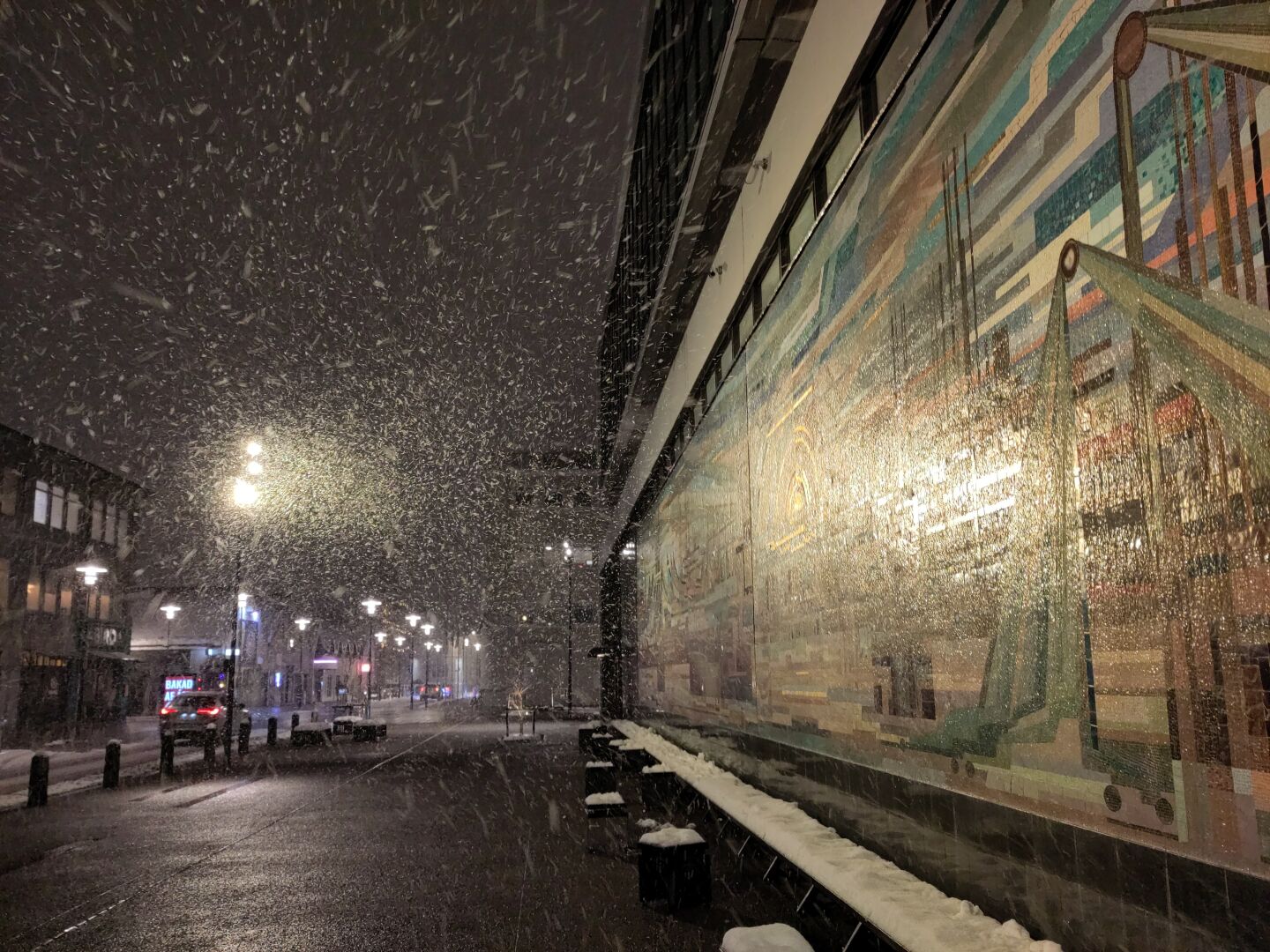 Night, heavy snow. A street with a wide sidewalk, lit by streetlamps. A side of a large building takes most of the shot. The wall is largely covered in a huge colorful relief / mosaic, showing harbor cranes and workers and other sea trade related things.