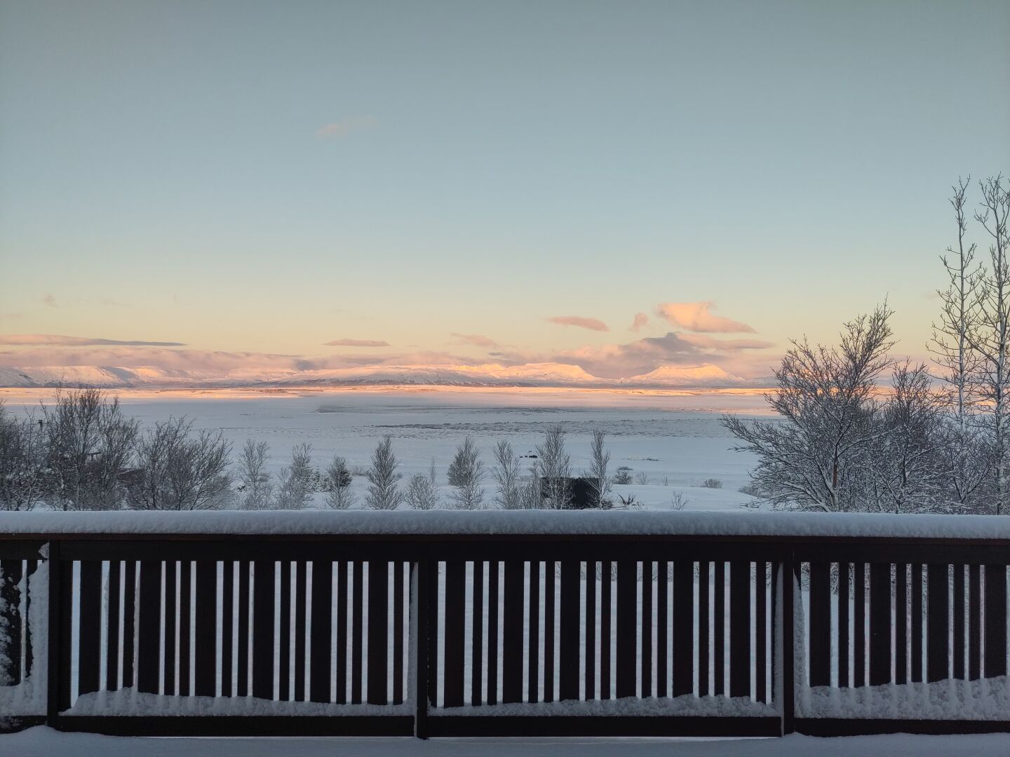 A calm winter afternoon over snowy fields, with mountains in the distance. Sky is a gradient from lilac to cool blue, some sparse clouds.